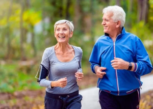 Two people with white hair and wrinkles on their faces jog outdoors while smiling.