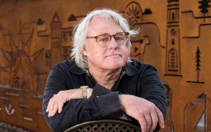 Man with white hair sitting backwards on a chair in front of a brown mural